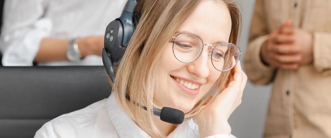 A smiling woman wearing round glasses and a headset with a microphone looks slightly downward while adjusting the earpiece, with two blurred coworkers in the background.