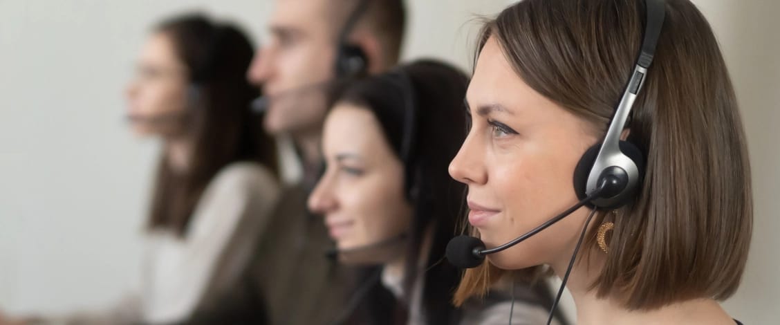 Four customer service representatives sit in a row wearing headsets with microphones. The woman in the foreground is in sharp focus, while the other agents are softly blurred in the background.