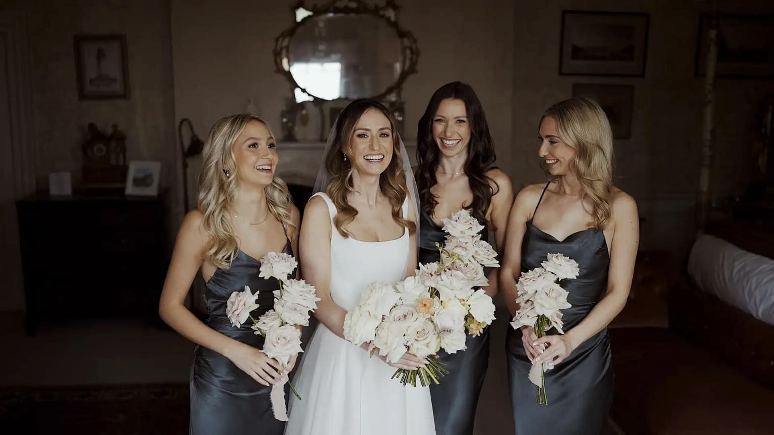 Bride with bridesmaids, holding bouquets, smiling indoors.
