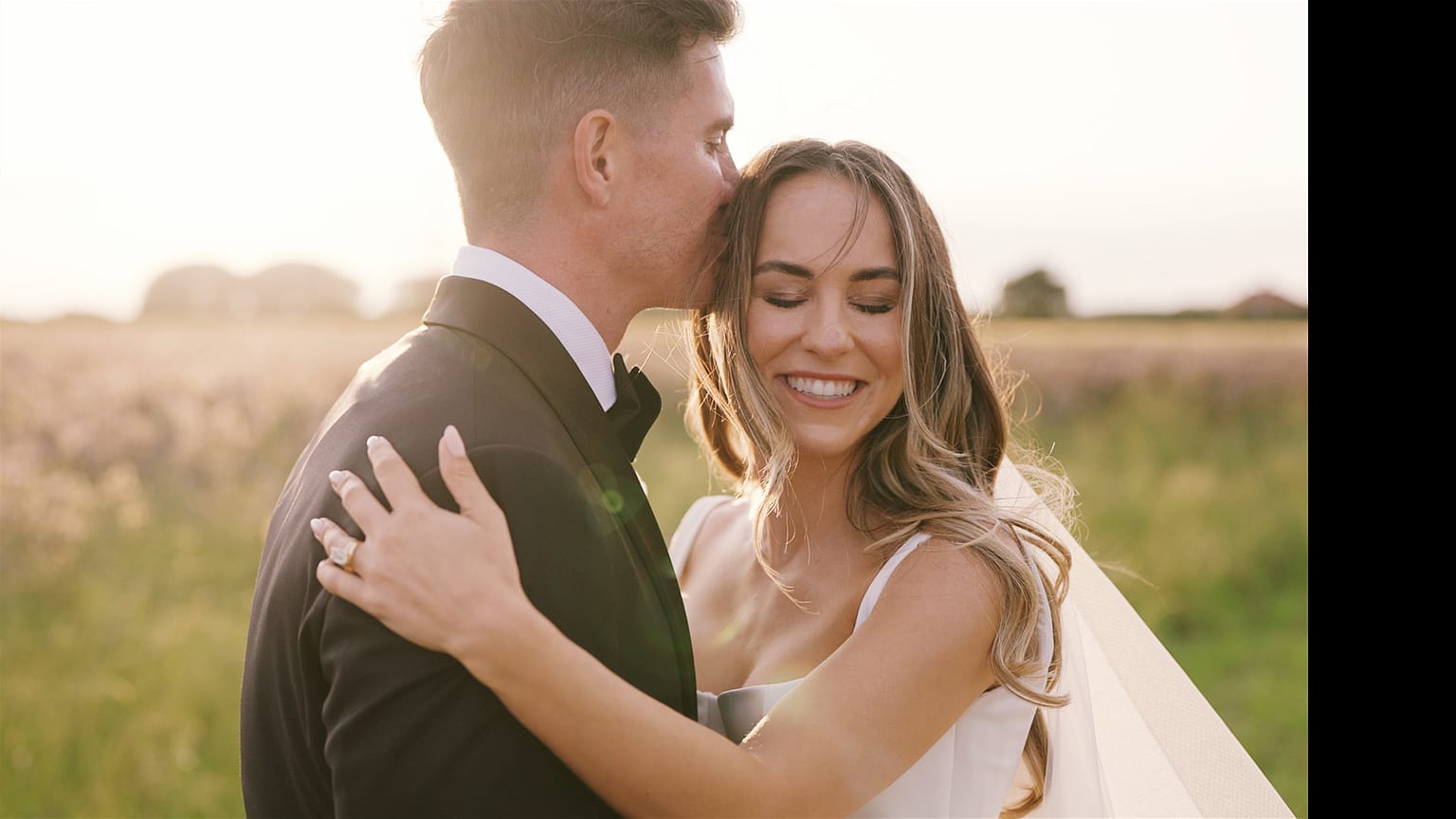 Bride and groom embrace in sunlit field.
