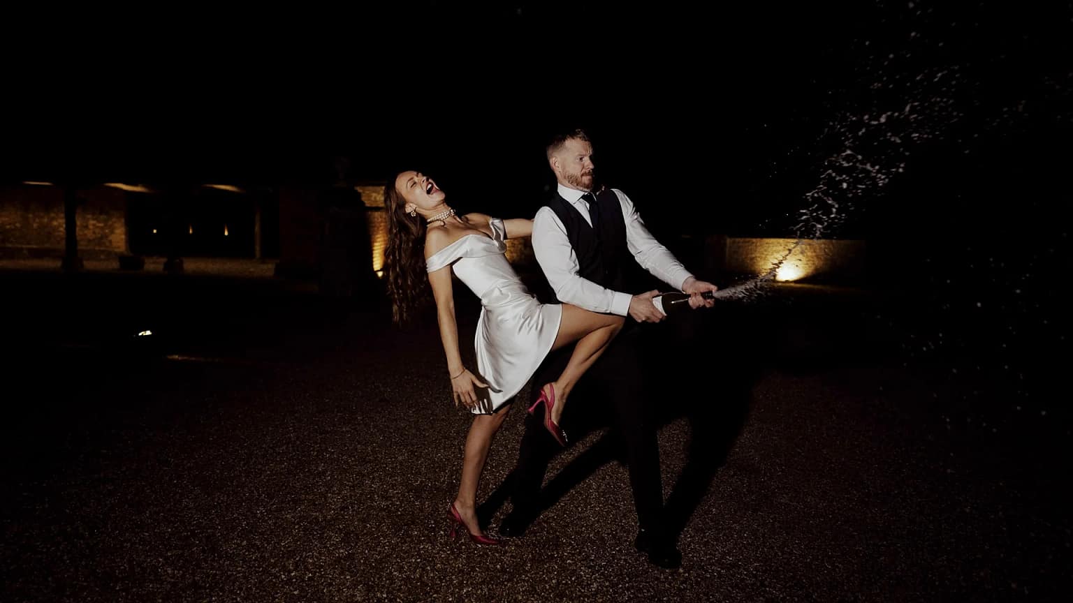 Couple joyfully spraying champagne at night celebration.