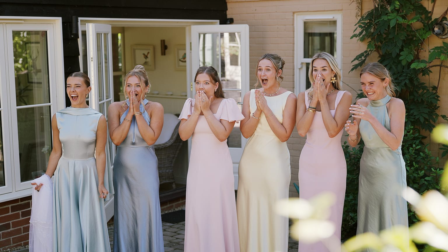 Bridesmaids in pastel dresses reacting with surprise outdoors