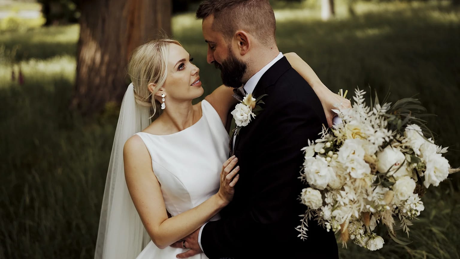 Bride and groom embrace with bouquet outdoors.