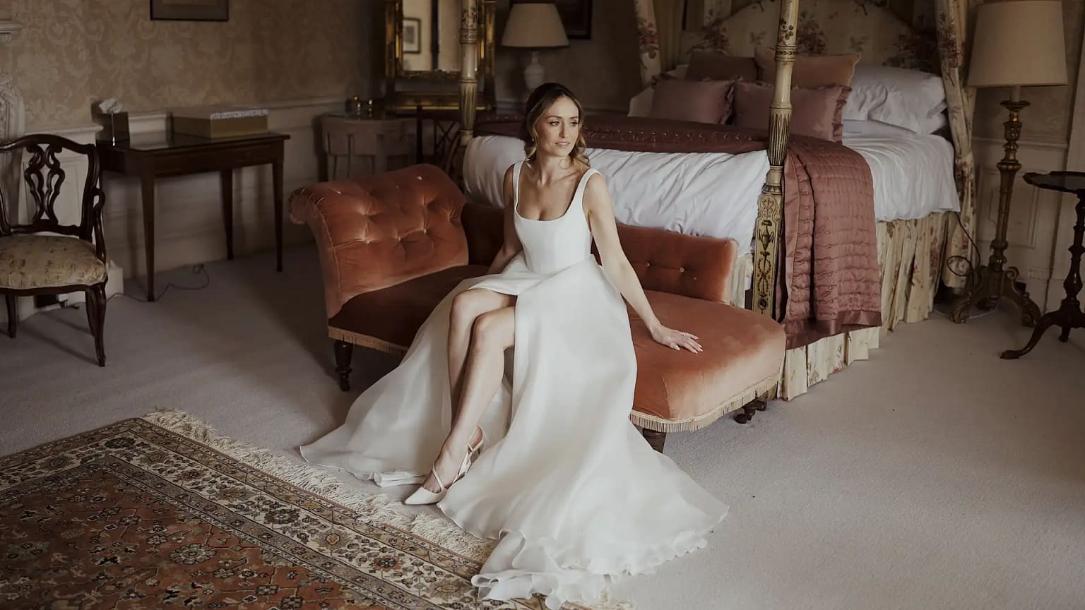 Bride sitting in elegant bedroom wearing white gown.