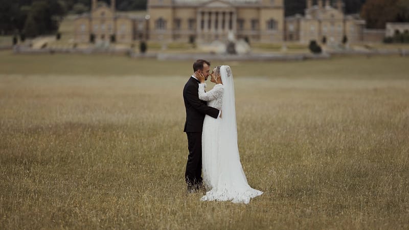 Bride and groom embracing in field, historic building backdrop.